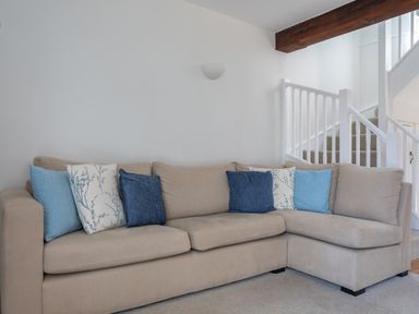 A beige L-shaped sofa with blue and patterned cushions in a living room near a staircase with white railing and a wooden beam at Stable End Cottage in Malborough