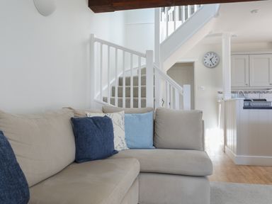 A living room with a beige sectional sofa with blue and white cushions near a white staircase and kitchen at Stable End Cottage in Malborough