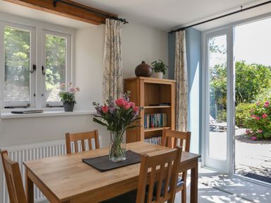 A dining area with a wooden table and four chairs a vase with flowers on the table a bookcase with books and plants curtains on windows and glass doors leading outside at Stable End Cottage Malborough