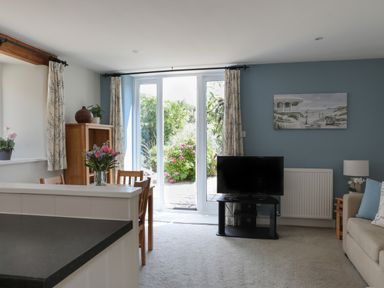 A living room with a television, dining table with flowers, and glass doors opening to a garden at Stable End Cottage in Malborough