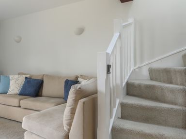 A beige sectional sofa with blue and patterned cushions next to a carpeted staircase with white railing at Stable End Cottage in Malborough