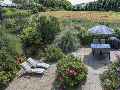 An outdoor garden area with two lounge chairs on gravel and a patio with table chairs and umbrella at Stable End Cottage in Malborough