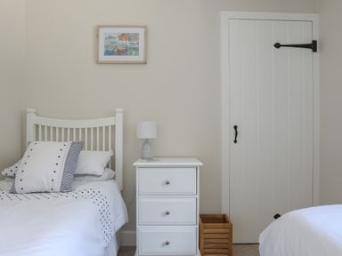 A bedroom with two single beds a white nightstand with a lamp a small wooden basket and a closed white door at Stable End Cottage in Malborough