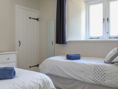 A bedroom with two single beds a window with dark curtains a white door and a white drawer at Stable End Cottage in Malborough