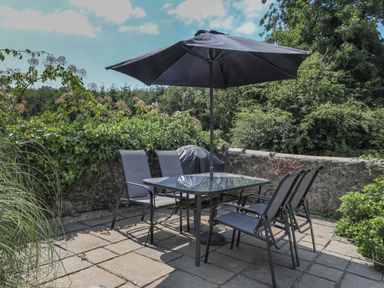 An outdoor patio with a glass table and four chairs under a large umbrella at Stable End Cottage in Malborough