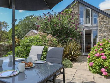 An outdoor patio area with a glass table and chairs set with plates and cups near a stone cottage with open doors surrounded by plants and flowers at Stable End Cottage in Malborough