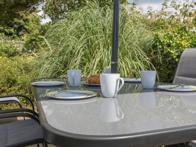 An outdoor glass table set with plates cups and croissants surrounded by chairs and plants at Stable End Cottage in Malborough