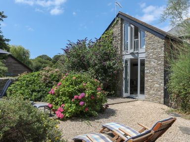 An outdoor patio area with two wooden lounge chairs and flowering shrubs outside a stone house at Stable End Cottage in Malborough
