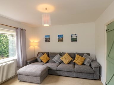 A living room with a grey sectional sofa and yellow and grey cushions near a window with curtains and a floor lamp at 7 Dale End in Danby near Castleton