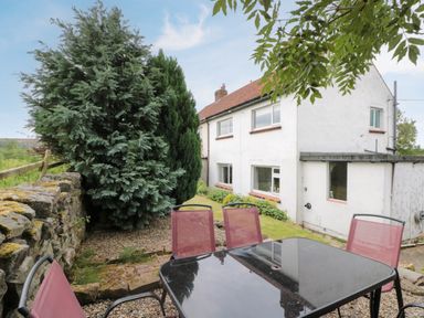 A garden patio with a black glass table and four red chairs beside a white house with large trees and a stone wall at 7 Dale End in Danby near Castleton