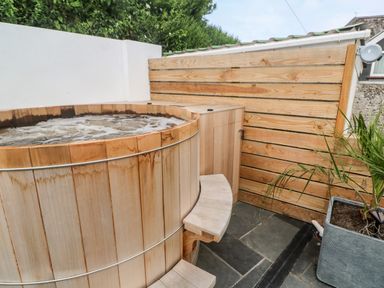 An outdoor wooden hot tub with bubbling water next to a wooden privacy fence and a potted plant at Postcard Lodge in Polruan