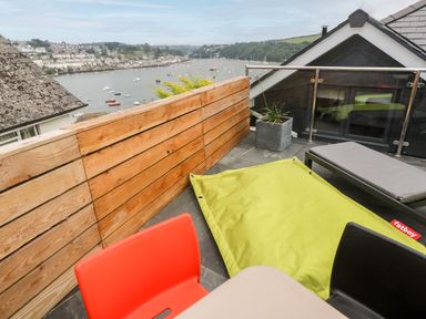 A rooftop terrace with a wooden fence, red and black chairs, a lime green bean bag, a gray lounge chair, and a view of a harbor with boats at Postcard Lodge in Polruan