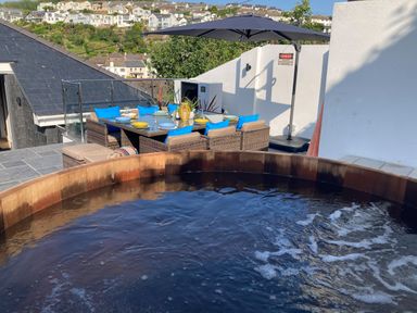 An outdoor patio with a bubbling wooden hot tub a dining table set with plates and blue cushions and an umbrella at Postcard Lodge in Polruan