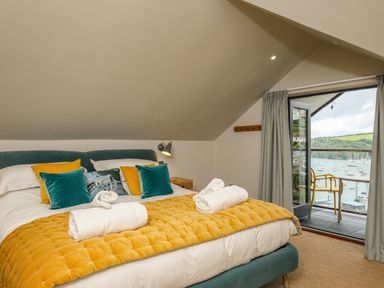 A bedroom with a double bed and pillows and an open door leading to a balcony with chairs overlooking a marina at Postcard Lodge in Polruan