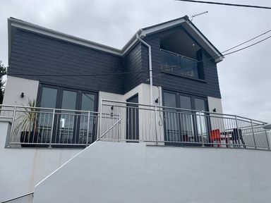 The exterior of a two-story house with dark upper siding and white lower walls with metal railings on the balcony at Postcard Lodge in Polruan