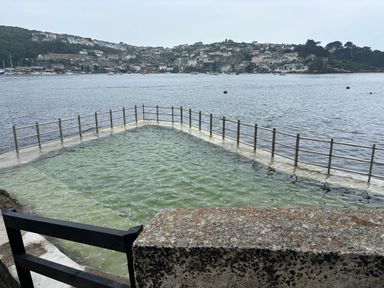 A seawater swimming pool with metal railings overlooking a body of water and a hillside with houses at Postcard Lodge in Polruan