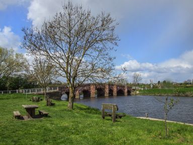 A park area with trees benches and picnic tables next to a river with a stone bridge in the background at The Hideaway in Eckington