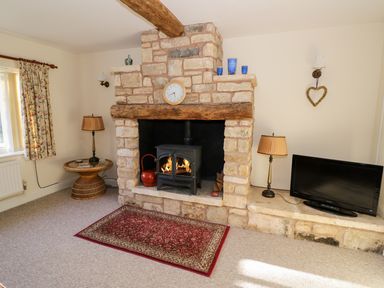 A living room with a stone fireplace with a wood stove and clock above it a small table with a lamp and floral curtains and a television on the hearth at The Hideaway in Eckington