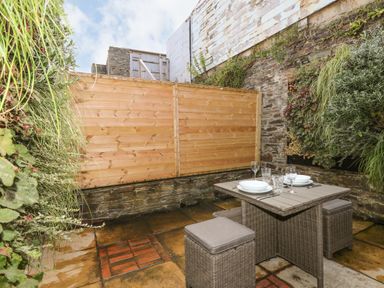 An outdoor patio area with a table set for two and two cushioned stools surrounded by stone walls and wooden fencing at Rock House Trebarwith Strand near Tintagel