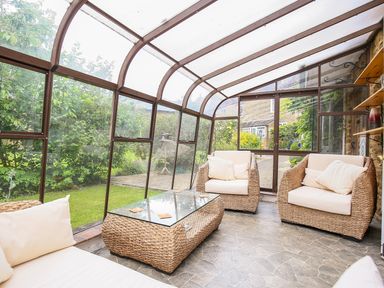 A conservatory with wicker armchairs and a glass coffee table overlooking a garden at Stone Wheel Cottage in Hook Norton