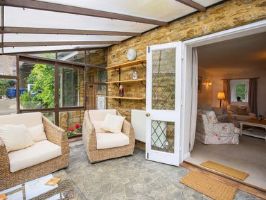 A sunroom with wicker armchairs and cushions next to a doorway leading to a living room at Stone Wheel Cottage in Hook Norton