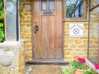 A wooden door with glass panels and a black knocker on a stone wall with a window and flowers in a pot at Stone Wheel Cottage in Hook Norton