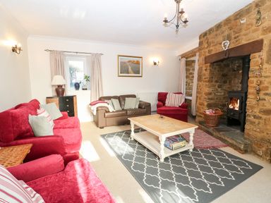 A living room with red and brown sofas a wooden coffee table a stone fireplace and a patterned rug at Stone Wheel Cottage in Hook Norton