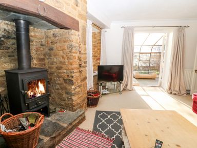 A living room with a wood-burning stove next to a stone wall a TV on a glass stand a wooden coffee table and an open door leading outside at Stone Wheel Cottage in Hook Norton