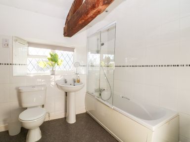 A bathroom with a toilet, pedestal sink, bathtub with shower, a window with plants and wooden beam at Stone Wheel Cottage in Hook Norton