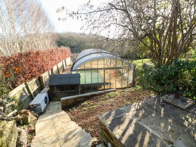 An outdoor area with stone steps leading down to a large greenhouse and a wooden shed surrounded by trees and shrubs at Stone Wheel Cottage in Hook Norton