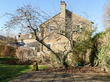A stone house with a glass conservatory and leafless trees in a garden at Stone Wheel Cottage in Hook Norton