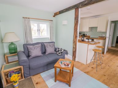 A living room with a blue sofa, wooden side table, green lamp, small wooden coffee table, and an open kitchen with two stools at Wigrams Canalside Cottage in Napton-on-the-Hill