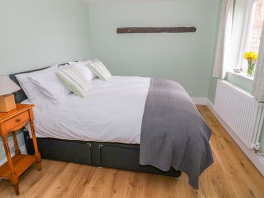A bedroom with a bed with white and striped pillows bedside table with a lamp wooden floor and a window with flowers at Wigrams Canalside Cottage in Napton-on-the-Hill