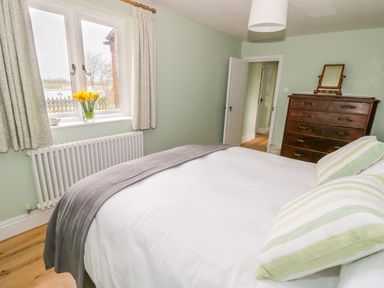 A bedroom with a bed with striped pillows a wooden dresser with a mirror and a window with curtains and flowers at Wigrams Canalside Cottage in Napton-on-the-Hill