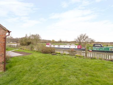 A grassy garden with a fence overlooking a canal with narrowboats near a brick house at Wigrams Canalside Cottage in Napton-on-the-Hill