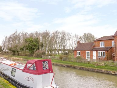 A narrowboat on a canal beside a brick cottage with a fenced garden and leafless trees in the background at Wigrams Canalside Cottage in Napton-on-the-Hill