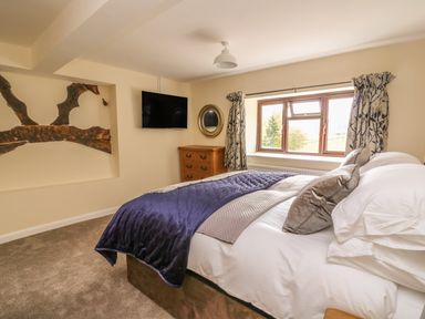 A bedroom with a bed pillows and blanket a window with curtains a wooden chest of drawers and a wall mounted television at Treboeth in Knighton