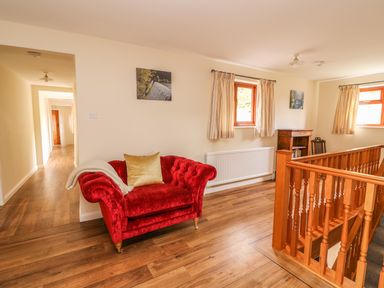 A seating area with a red armchair a wooden stair railing two windows with curtains and a small bookshelf at Treboeth in Knighton