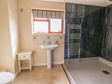 A bathroom with a pedestal sink a small white side table a window with striped blinds and a walk-in shower with dark wall panels at Treboeth in Knighton