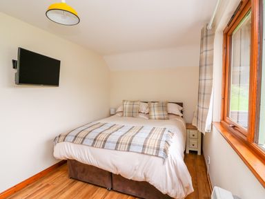 A bedroom with a double bed pillows bedside tables a wall mounted tv and a window with curtains at Pines Pitch Knighton