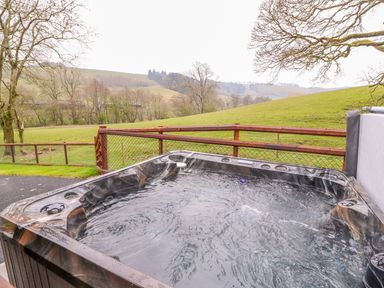 An outdoor hot tub with bubbling water and a wooden fence overlooking a grassy hill and trees at Pines Pitch in Knighton