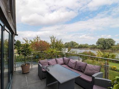 An outdoor balcony with a wicker sofa set and glass table overlooking a river and trees at Longmynd in Gloucester