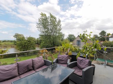 An outdoor seating area with a glass table and cushioned chairs overlooking a garden river and trees at Longmynd in Gloucester