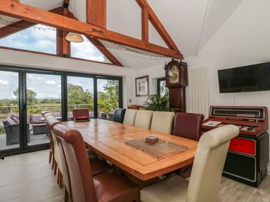 A dining room with a large wooden table surrounded by mixed color chairs and a grandfather clock beside a wall mounted television at Longmynd in Gloucester