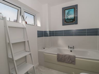 A bathroom with a bathtub with a towel hanging on it a white ladder shelf a framed picture on the wall and small windows above at Longmynd in Gloucester