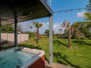 A covered patio with a hot tub overlooking a lawn with palm trees at Longmynd in Gloucester
