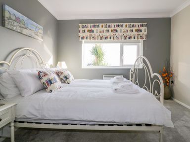 A bedroom with a white metal bed frame white bedding pillows with house patterns a window with patterned blinds a bedside table with a lamp and a vase with orange flowers at Bayview in Mevagissey