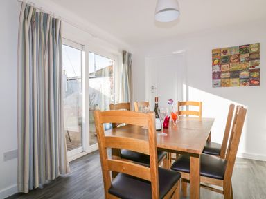 A dining room with a wooden table and six chairs near glass sliding doors with striped curtains and a colorful wall art at Bayview Mevagissey