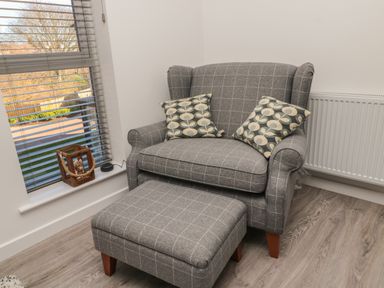 A corner with a gray checkered armchair and matching footstool with patterned cushions near a window with blinds at High Tide House in Craster