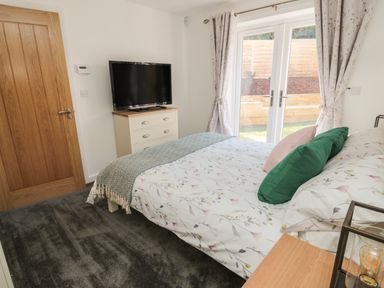 A bedroom with a bed with floral bedding two green and one pink pillow a chest of drawers with a television floral curtains and glass doors at High Tide House in Craster